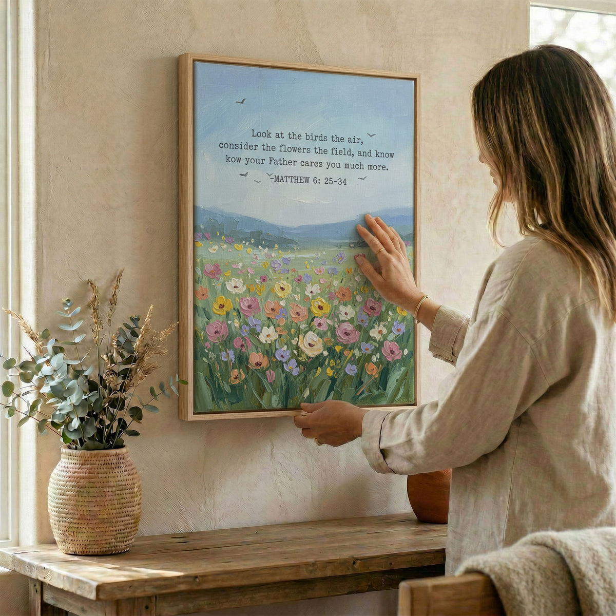 Woman hanging Bible verse wall art in wood frame beside dried botanical arrangement
