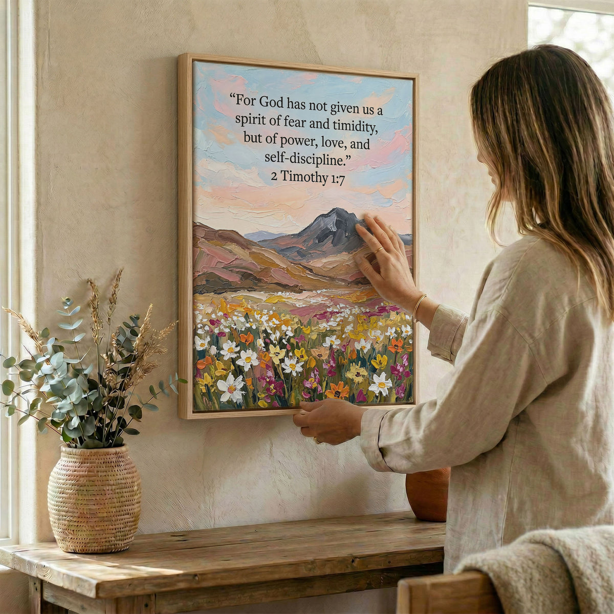 Woman hanging Bible verse wall art in wood frame beside dried botanical arrangement
