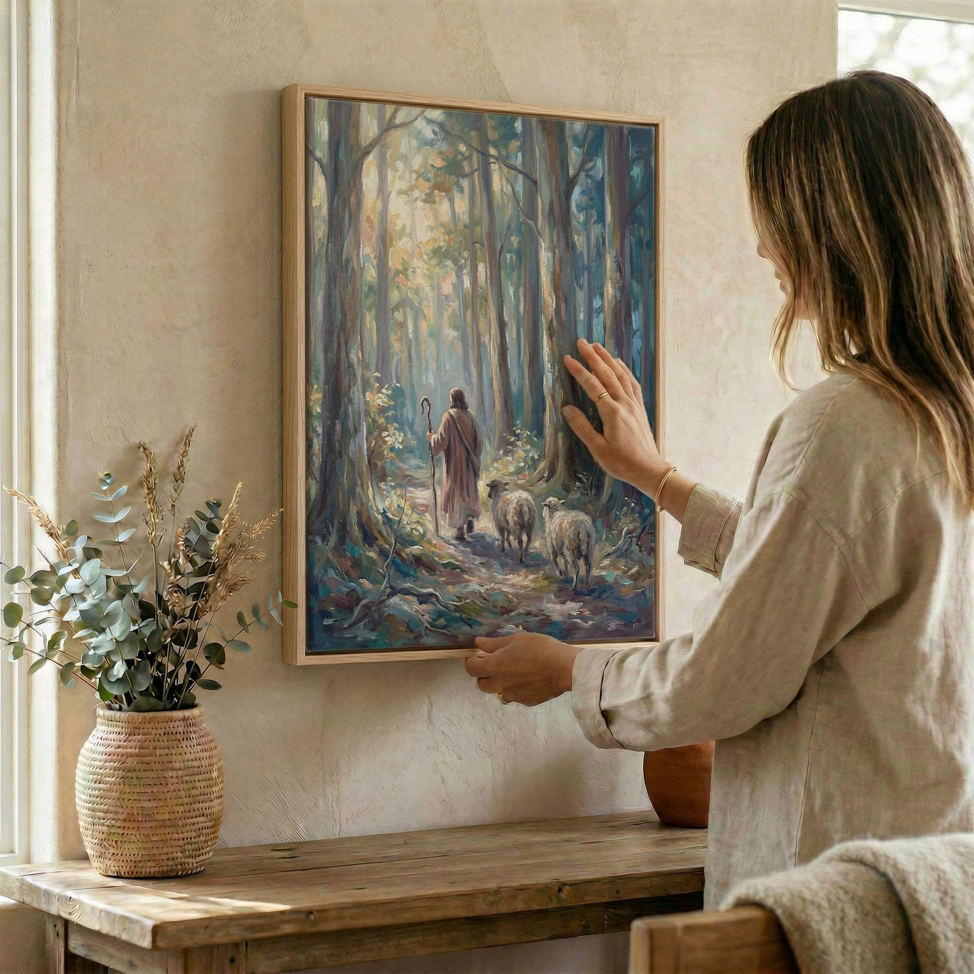 Woman hanging Good Shepherd sunlit forest wall art in wood frame beside dried botanical arrangement