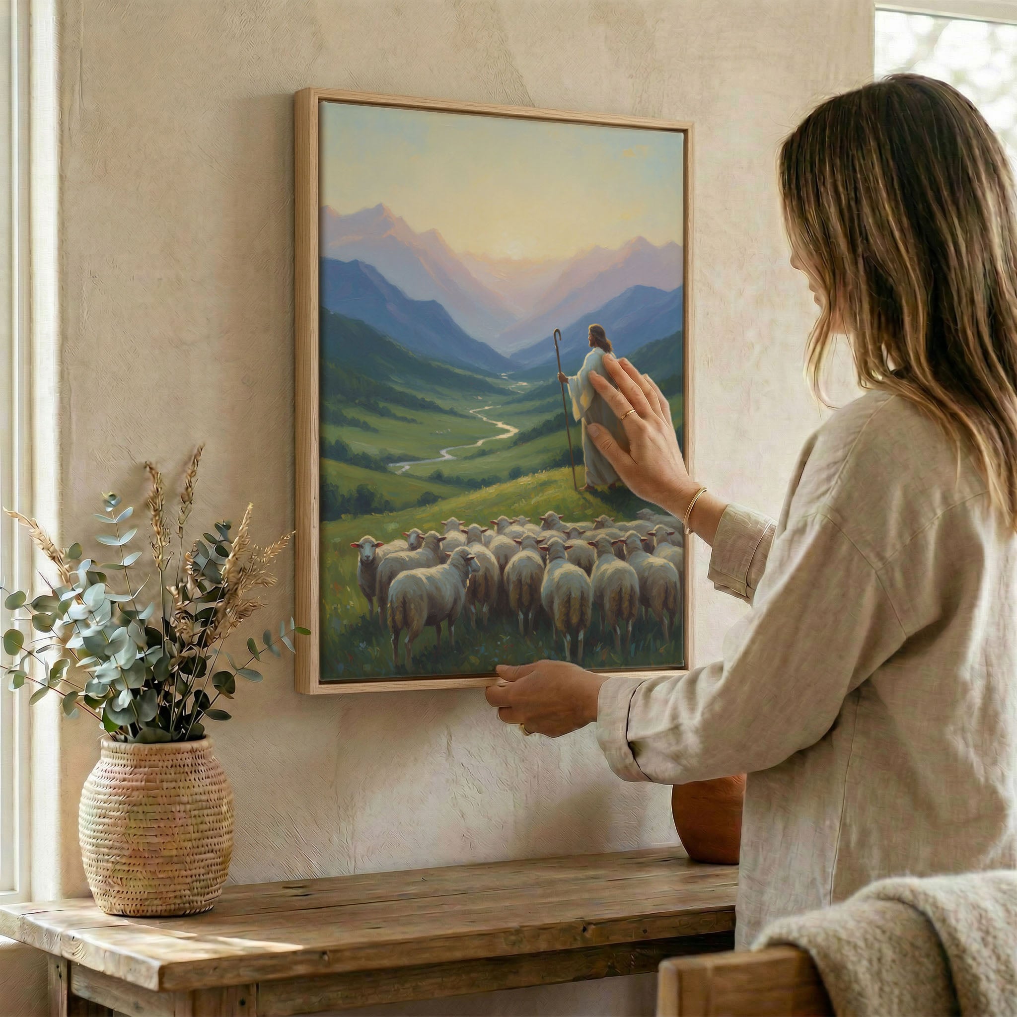 Woman placing Jesus Good Shepherd valley wall art in wood frame on wall beside dried botanical arrangement