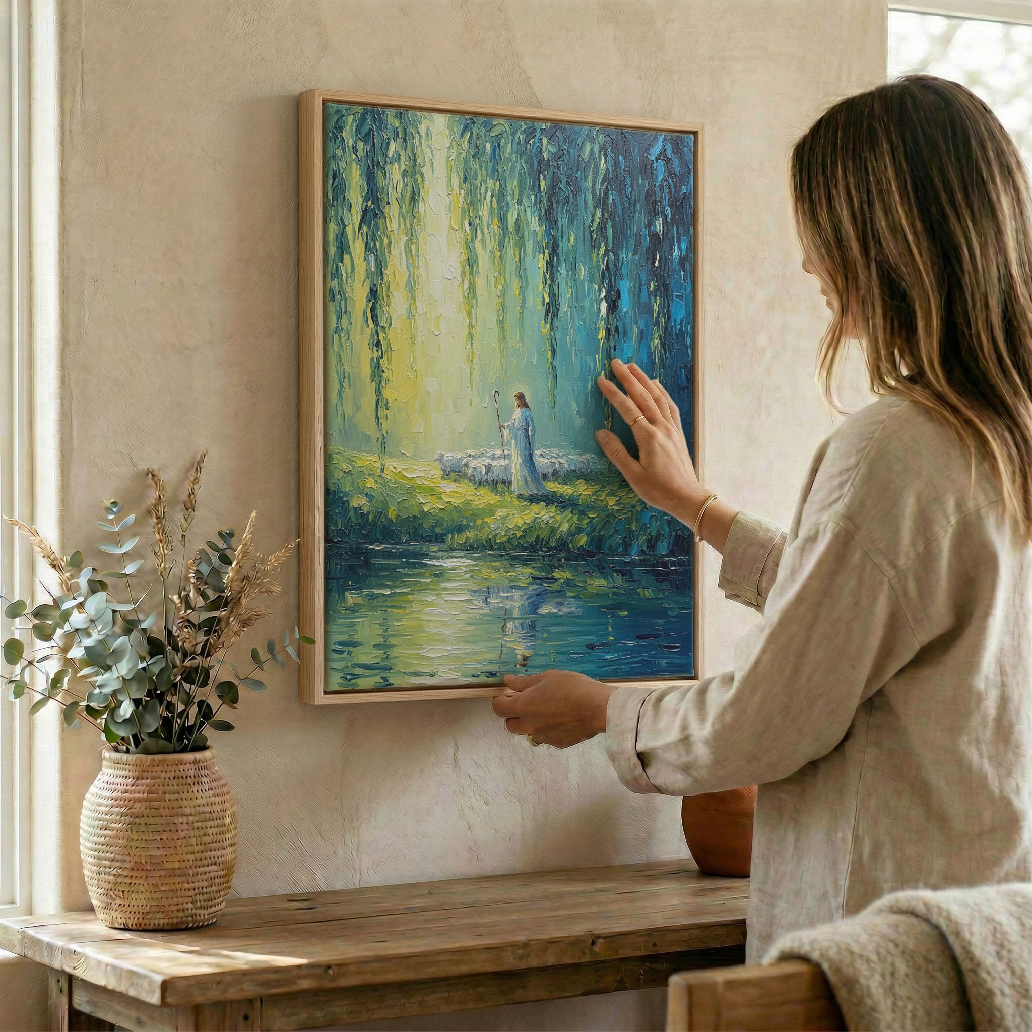 Woman placing Jesus Good Shepherd willow river wall art in wood frame on wall beside dried botanical arrangement