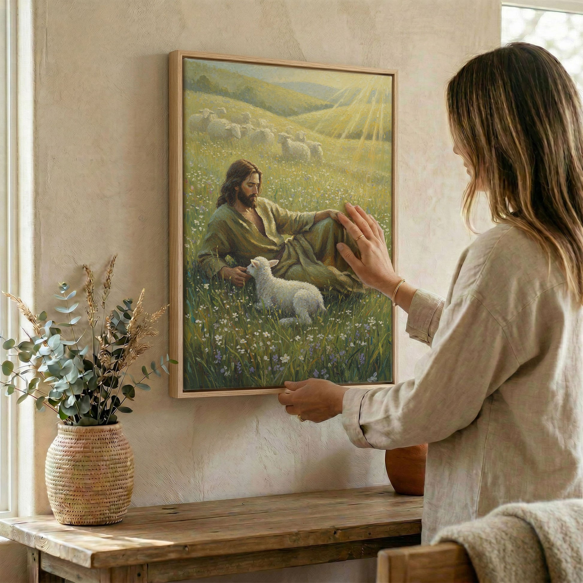 Woman placing Jesus Good Shepherd and lamb religious wall art in wood frame on wall beside dried botanical arrangement