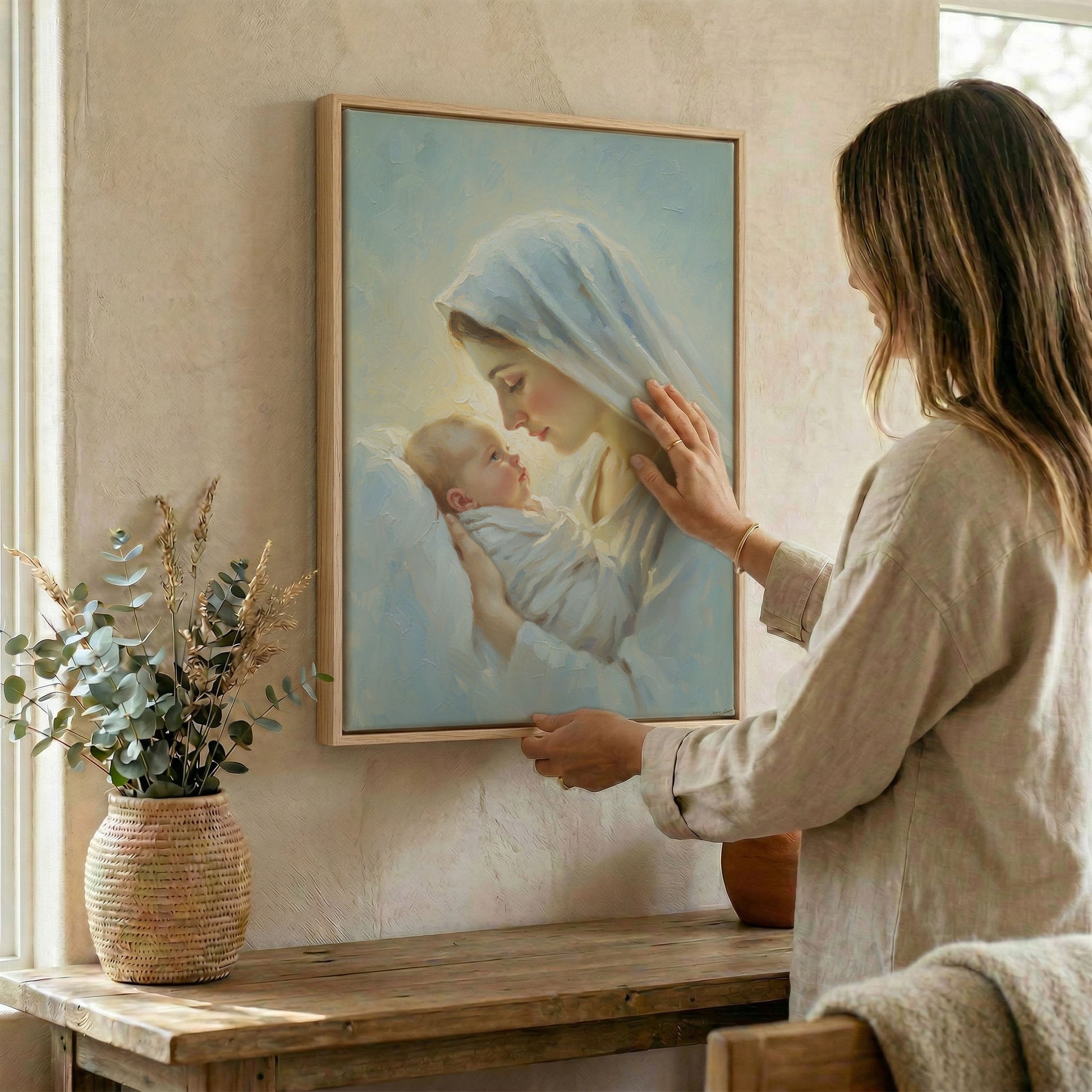 Woman placing Virgin Mary and Baby Jesus religious wall art in wood frame on wall beside dried botanical 