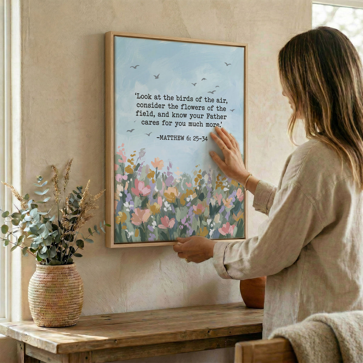 Woman placing Matthew 6:25-34 floral Christian wall art in wood frame on wall beside wicker basket arrangement